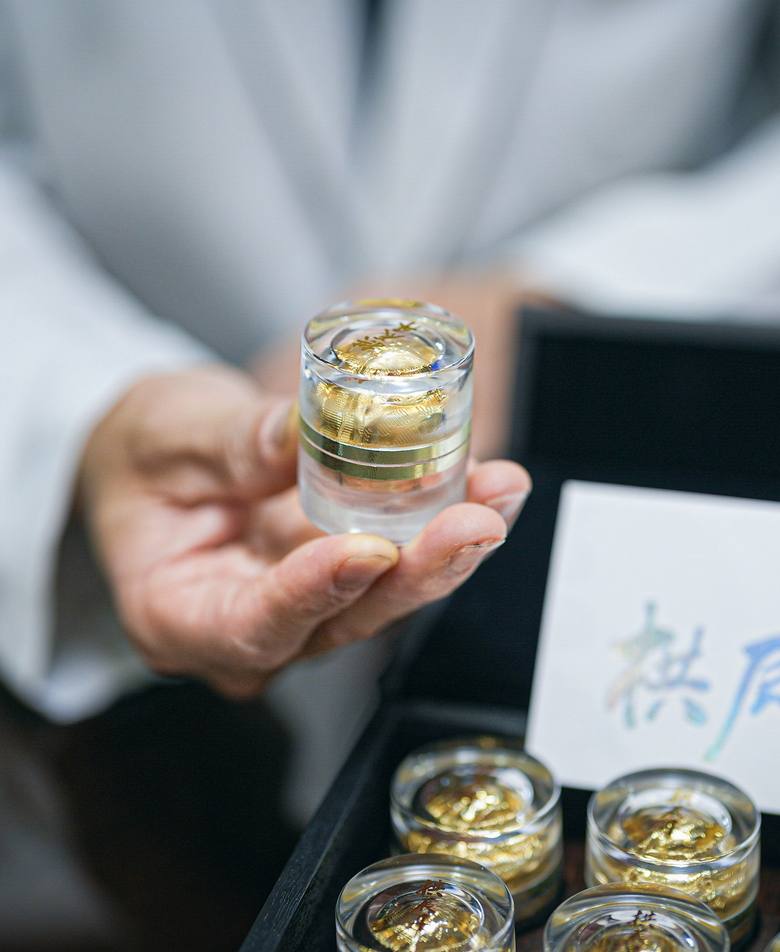 흰 가운을 입은 손에 금박 환이 담긴 투명 케이스를 올려 보여 주는 모습, 뒤로 케이스가 담긴 상자